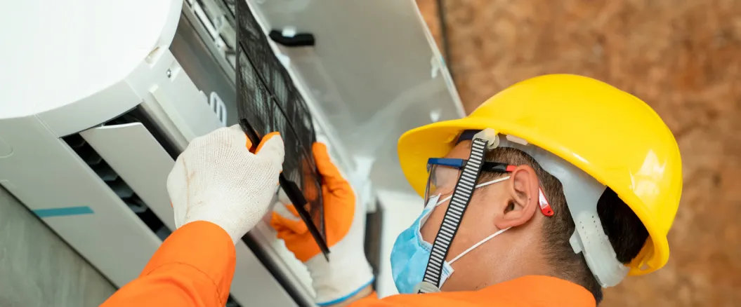 Technician wearing a yellow hard hat and protective gloves cleaning an air conditioner filter during maintenance service.