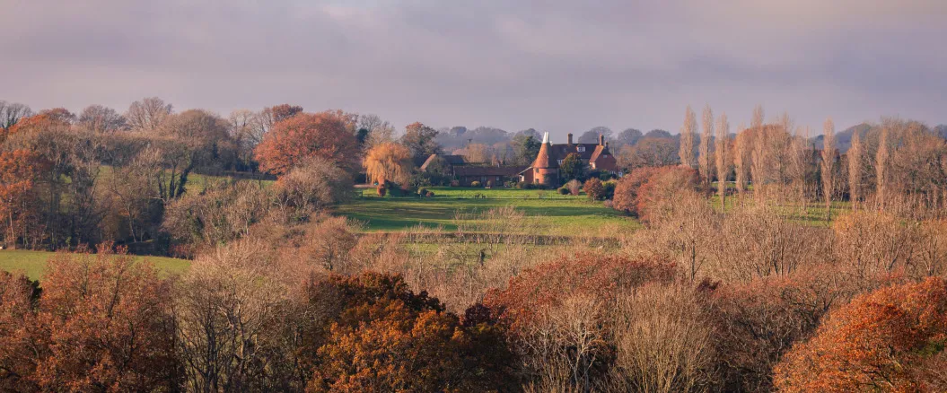 Scenic countryside landscape with a traditional red brick house surrounded by trees and green fields during autumn.