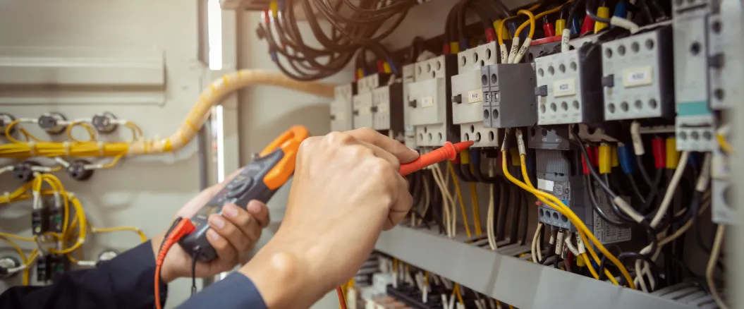 Electrician using a multimeter to test and troubleshoot electrical components in an industrial control panel with wires and switches.