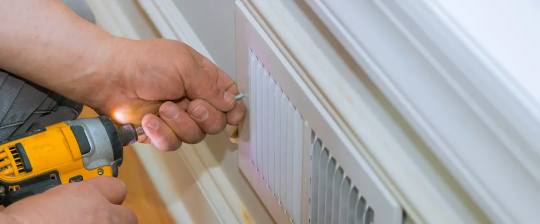 Technician using a power drill to secure or repair a white HVAC air vent cover on a residential wall."