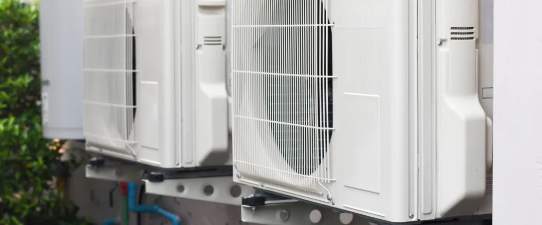 Two outdoor air conditioning units mounted on a wall with visible pipes and electrical connections.