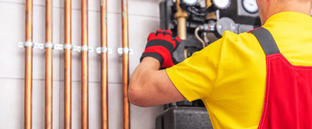 Technician in a yellow and red uniform working on a heating or plumbing system with copper pipes and gauges mounted on a wall.