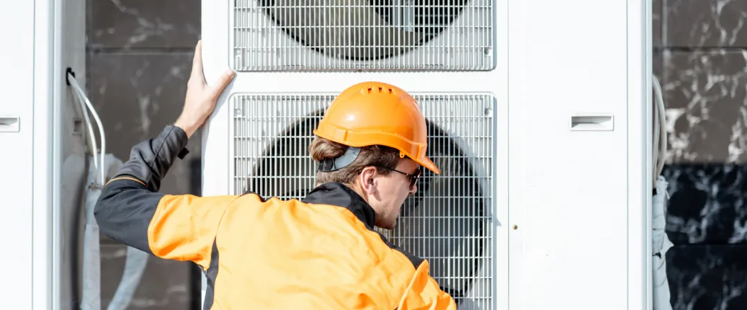 Technician in an orange hard hat and jacket inspecting a large industrial air conditioning unit.
