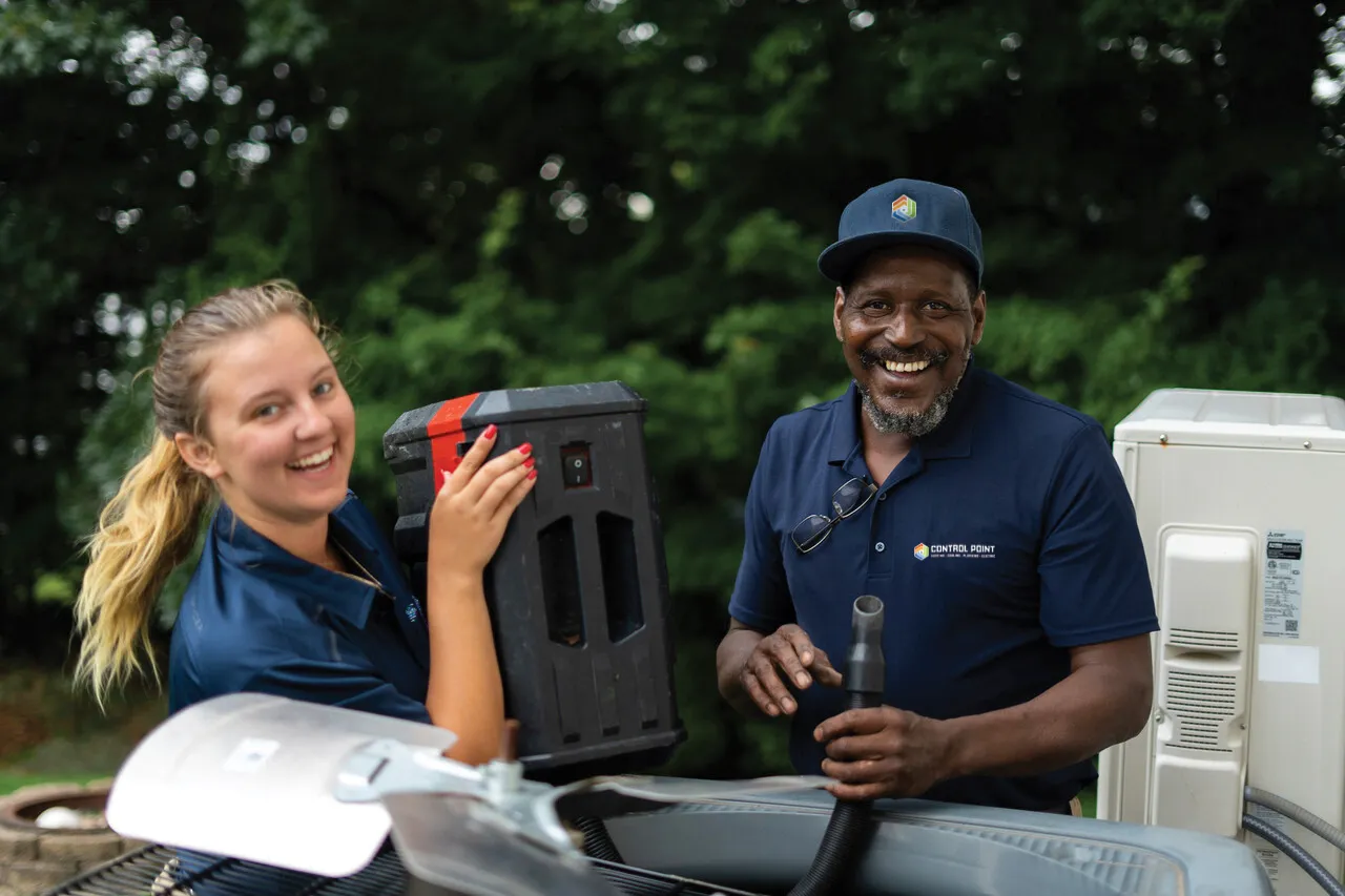Two HVAC technicians smiling while working on an outdoor air conditioning unit, with tools and equipment nearby.