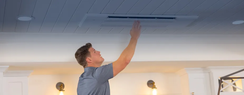 Man in a gray shirt reaching up to adjust a ceiling-mounted air conditioning vent.